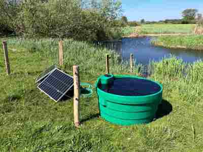 Soldriven markpump med solpanel och vattentråg vid en bäck på en äng.