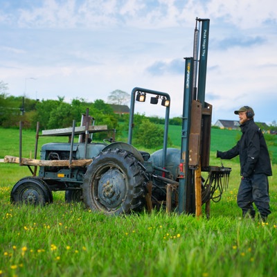 Montör arbetar med stolphammare på traktor vid uppsättning av stängselstolpar i ett grönt fältområde.